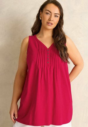 Kobieta o długich brązowych włosach nosi czerwoną tunikę bez rękawów z guzikami, stoi na jasnym, jednolitym tle.