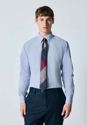 Young man wearing a light blue striped dress shirt, multicolor diagonal striped tie, and dark blue trousers, standing against a plain background.
