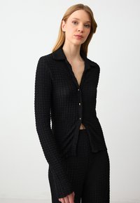 Zwarte button-up shirt van een textuurstof met een gekreukte afwerking. Heeft een kraag, lange mouwen en gouden knopen. Strakke pasvorm.