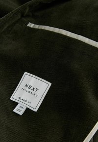 Tmavohnedá kordová látka s bielou etiketou s nápisom "NEXT TAILORING RELAXED FIT." Obsahuje viditeľnú textúru a kontrastné biele švy.