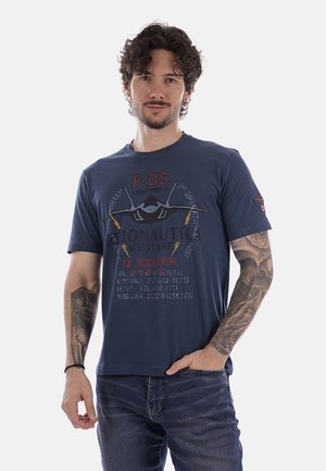 Uomo con capelli ricci e tatuaggi su entrambe le braccia indossa una maglietta blu navy con grafica del caccia F-35 e jeans scuri, in piedi con una mano in tasca.