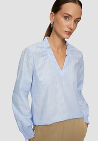 Blouse rayée bleu clair avec un col en V et un col volanté, dotée de manches longues avec des poignets froncés. Texture de tissu douce et lisse.