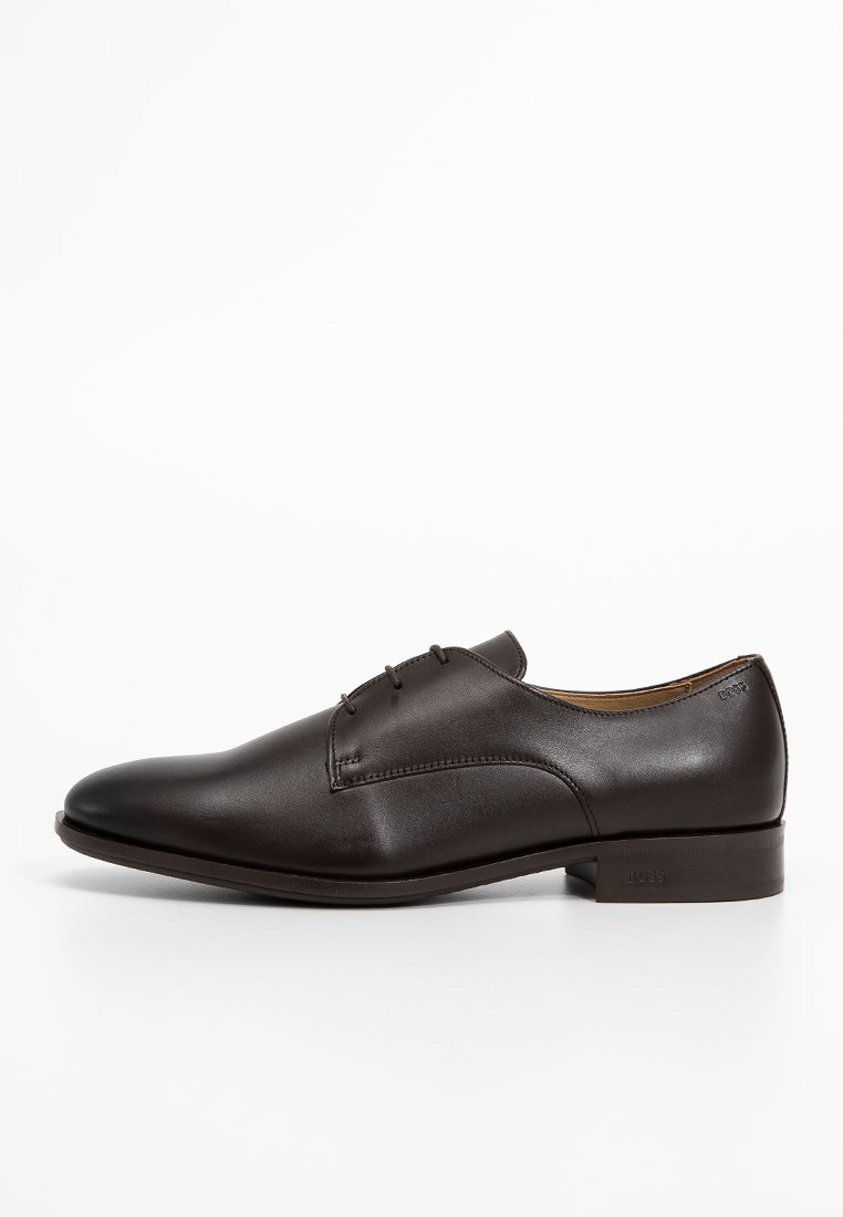 Chaussure de ville en cuir brun avec une texture lisse, bout rond, couture minimale et lacets étroits, arborant un petit logo de marque sur le côté.