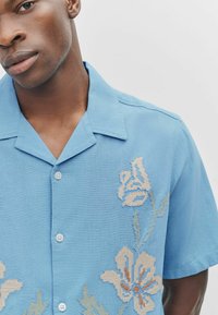 Kurzarmhemd in Hellblau mit floralen Stickereien in Beige und Grün, mit durchgehender Knopfleiste und gespreiztem Kragen.