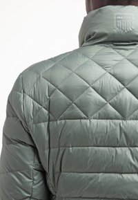 Quiltet grøn pufferjakke med høj krave, der har et diskret logo ved halsen og en skinnende, glat overflade.