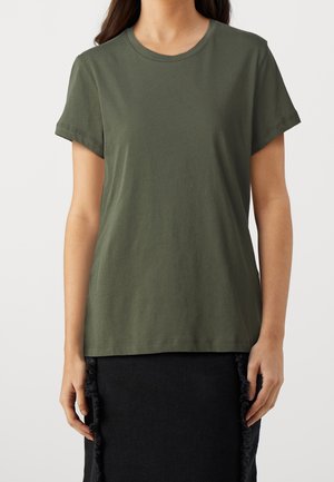 Person wearing plain dark green short-sleeve T-shirt and black skirt with frayed side details, standing against a plain light background.