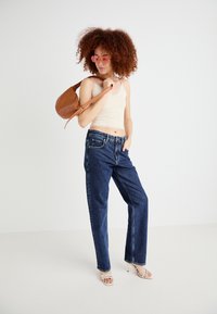 Vaqueros de mezclilla en azul oscuro, de corte holgado con diseño de cinco bolsillos. Combinados con una camiseta sin mangas beige claro y un bolso de hombro marrón. Gafas de sol transparentes de color rosa.