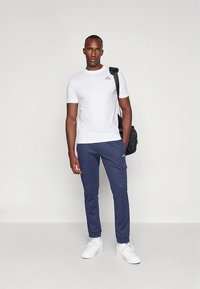 Λευκό t-shirt με στρογγυλή λαιμόκοψη και μικρό λογότυπο, συνδυασμένο με μπλε ναυτικό cargo παντελόνι με τσέπες. Παπούτσια λευκά αθλητικά. Μαύρη τσάντα ώμου.