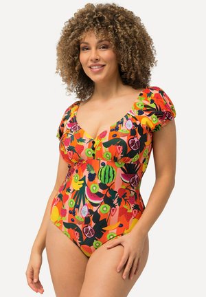 Kobieta o kręconych włosach, uśmiechająca się, ubrana w kolorowy jednoczęściowy strój kąpielowy z różnymi wzorami owoców, opierająca rękę na udzie.