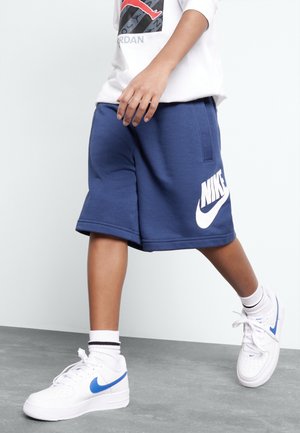Dziecko noszące niebieskie szorty Nike, białe buty Nike z niebieskim logo, białe skarpetki z czarnymi paskami, chodzące po szarej podłodze.