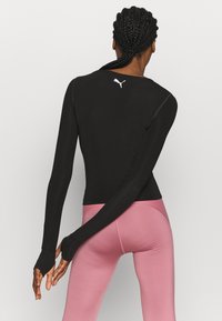 Czarna sportowa bluzka z długim rękawem o dopasowanym kroju, z otworami na kciuki przy mankietach i białym logo z tyłu, zestawiona z różowymi legginsami.