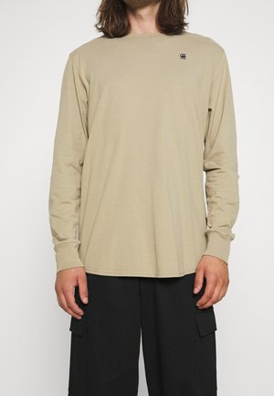 Homme portant une chemise beige à manches longues avec un petit logo sur la poitrine et un pantalon cargo noir, debout devant un fond uni.