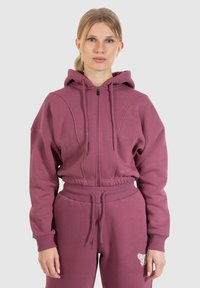 Felpa zip-up color bordeaux con cappuccio e polsini a costine, abbinata a pantaloni jogger coordinati con un dettagli ricamato. Tessuto morbido e con texture.