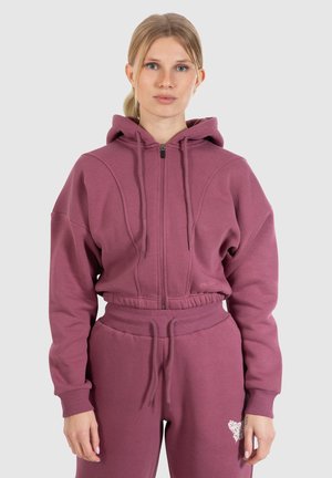 Haut zippé bordeaux avec capuche et poignets côtelés, associé à un pantalon de jogging assorti avec un détail brodé. Tissu doux et texturé.
