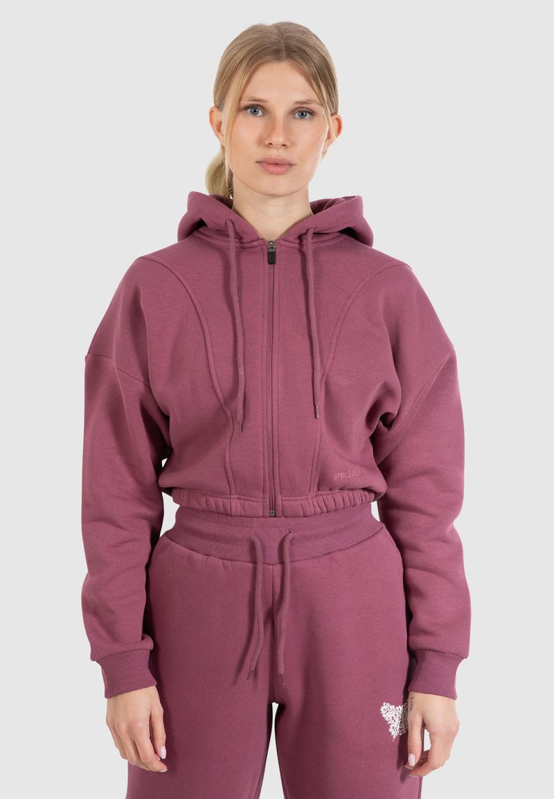 Felpa zip-up color bordeaux con cappuccio e polsini a costine, abbinata a pantaloni jogger coordinati con un dettagli ricamato. Tessuto morbido e con texture.