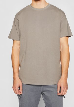 Mann in einem einfarbigen taupefarbenen T-Shirt mit Kurzarm und grauen Hosen mit Seitentaschen, steht mit entspannten Armen an den Seiten.