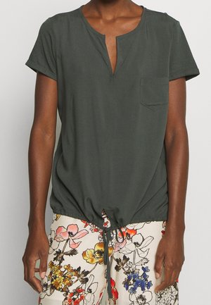 Persoon draagt een donkergroene korte mouw top met voorzak en trekkoord aan de zoom, gecombineerd met kleurrijke bloemenprint broek.