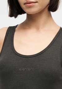 Gros plan d'une personne portant un débardeur côtelé foncé avec le mot « element » brodé sur la poitrine.