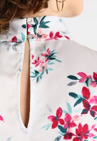 Blusa floreale realizzata in tessuto bianco con motivi rosa e verdi, colletto alto e chiusura a bottoncino sul retro con dettagli a oblò.