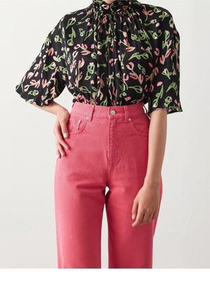 Kvinde iført lyserøde bukser med høj talje og en sort bluse med lyserøde og grønne blomsterprint, stående med den ene hånd på hoften.