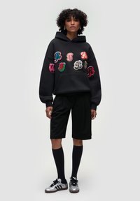 Felpa nera con lettere ricamate multicolori, abbinata a pantaloni corti neri, calze nere al ginocchio e sneaker argento con accenti neri.