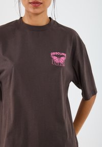Dark brown cotton T-shirt with short sleeves, featuring a pink butterfly graphic and the text "OBSCURE FUTURE" on the left chest area.