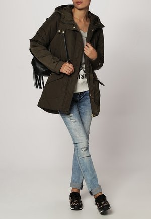 Khakifarbene gepolsterte Jacke mit Kapuze, ausgestattet mit großen Taschen und goldfarbenen Beschlägen. Kombiniert mit hellblauen, verwaschenen Jeans und gemusterten schwarzen Sneakers.