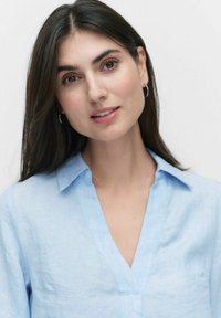 Ljust blå, linneskjorta med krage och v-ringning. Slät textur med subtila veck. Modellen har långt, mörkt hår och stora örhängen.