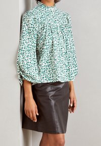 Blumenbluse in Grün und Weiß, mit hohem Ausschnitt und Puffärmeln. Kombiniert mit einem taillierten, glänzenden schwarzen Lederrock.