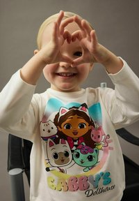 Uśmiechnięte dziecko w białej koszulce Gabby's Dollhouse, formujące kształt serca dłońmi przed twarzą na neutralnym tle.