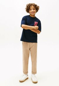 Marineblå t-shirt med et rødt "H" logo, parret med beige bukser med opfoldede ærmer og hvide sneakers med brune detaljer. Barnet står med armene krydset.