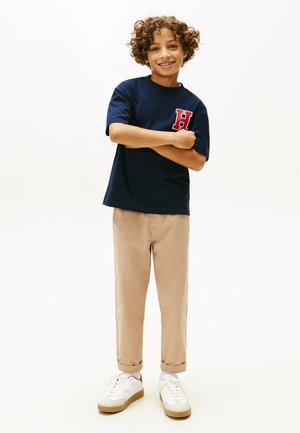 Navyblauwe t-shirt met een rood "H" logo, gecombineerd met beige broek met opgerolde manchetten en witte sneakers met tan accenten. Kind poseert met armen gekruist.
