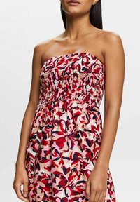 Frau in einem trägerlosen Kleid mit rotem, pinkem und schwarzem Blumenmuster und gerafftem Oberteil, die vor einem schlichten Hintergrund steht.