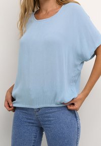 Hellblaues, kurzärmliges Oberteil aus plissiertem Stoff mit Rundhalsausschnitt und lässiger Passform, kombiniert mit dunkelblauen Jeans.