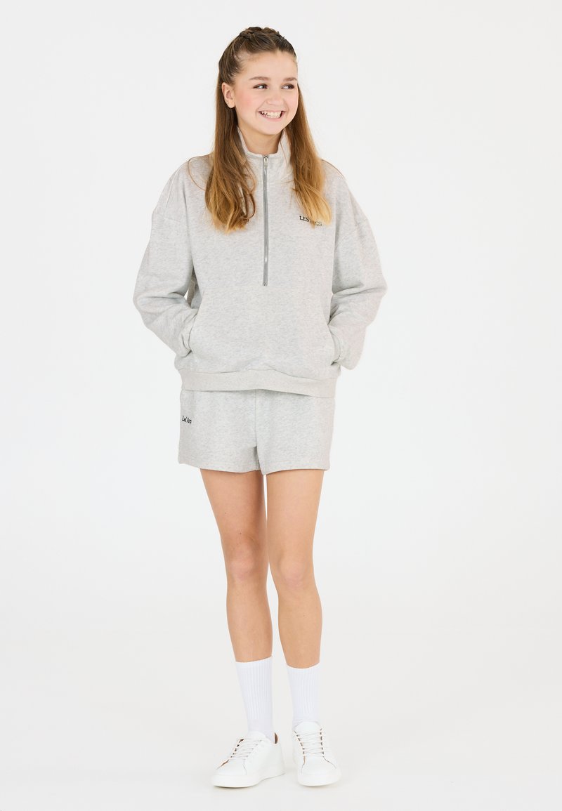 Grauer Fleece-Sweatshirt mit halbem Reißverschluss-Kragen und Fronttaschen, kombiniert mit passenden Shorts. Weiße Sneakers und knielange Socken runden den Look ab.