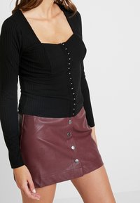 Mustapohjainen pitkähihainen toppi, jossa on neliön muotoinen pääntie ja keskellä napitus, yhdistettyään burgundinpunaisen tekonahkahameen, jossa on metalliset napit.