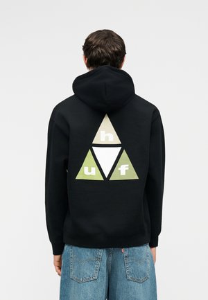 Osoba u crnoj majici s kapuljačom na leđima s geometrijskim trokutastim logom i slovima "h", "u", "f", u kombinaciji s plavim trapericama.