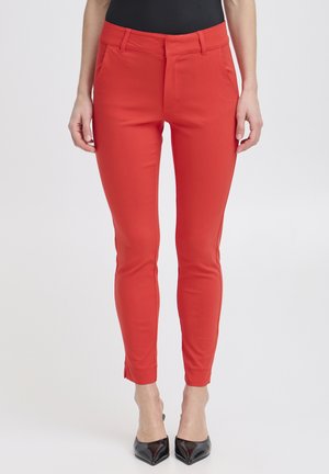 Personne portant un pantalon rouge slim et des escarpins noirs pointus, debout devant un fond clair uni, visible du buste aux pieds.