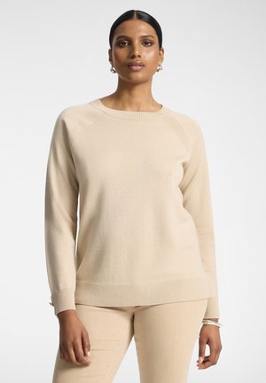 Donna con capelli scuri che indossa orecchini a cerchio dorati, maglione beige e pantaloni beige su sfondo bianco neutro.