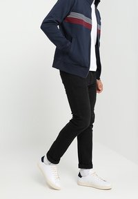 Giacca blu navy con zip, a righe orizzontali rosse e bianche, abbinata a jeans neri e sneaker bianche con accenti blu navy. Texture liscia.