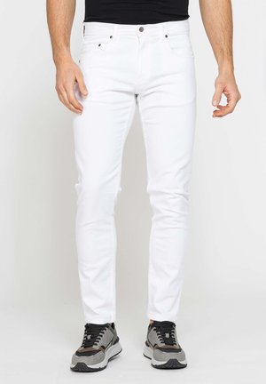 Uomo che indossa jeans bianchi slim fit con sneakers nere e grigie, in piedi davanti a uno sfondo chiaro uniforme.