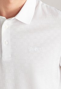 Bílá strukturovaná polo tričko s límečkem, dvěma knoflíky a jemně embosovaným logem "JOOP!" na hrudi, nošené osobou.