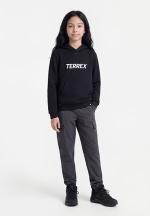 Unge person, der står iført en sort "TERREX" hættetrøje, mørkegrå cargobukser og sorte vandresko mod en ensfarvet hvid baggrund.