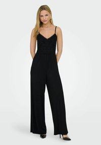 Combinaison noire avec un corsage en dentelle aux bords festonnés, fines bretelles, jambes larges et une texture de tissu lisse. Talons hauts noirs inclus.