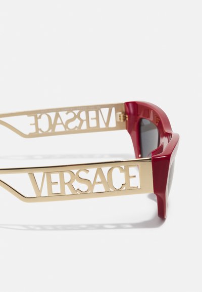 Punased plastist prillid julge geomeetrilise disainiga, millel on kuldmetallist sangad, kuhu on sisse lõigatud "VERSACE" tähed. Tumedad läätsed.