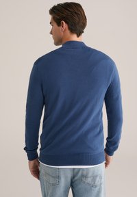 Pulover bărbați, de culoare albastră, din material tricotat, cu design de guler, mâneci lungi și manșete și tiv cu nervuri. Purtați peste o cămașă de culoare deschisă și blugi.