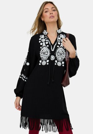 Frau trägt schwarze Langarm-Tunika mit weißer Blumenstickerei, Fransen am Saum, trägt eine braune Schultertasche vor einfachem Hintergrund.