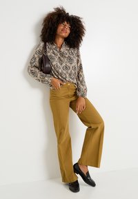 Blusa de estampado floral en beige y negro, combinada con pantalones anchos mostaza. Mocasines negros completan el look. Acento de bolso marrón.