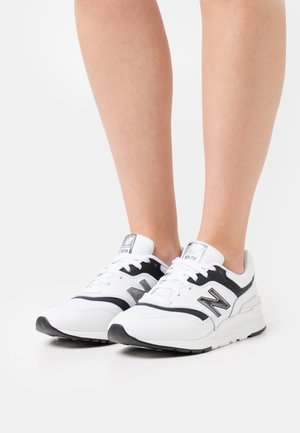 Osoba oblečená v bílých a černých teniskách New Balance 997H s tkaničkami, stojící na bílém pozadí.
