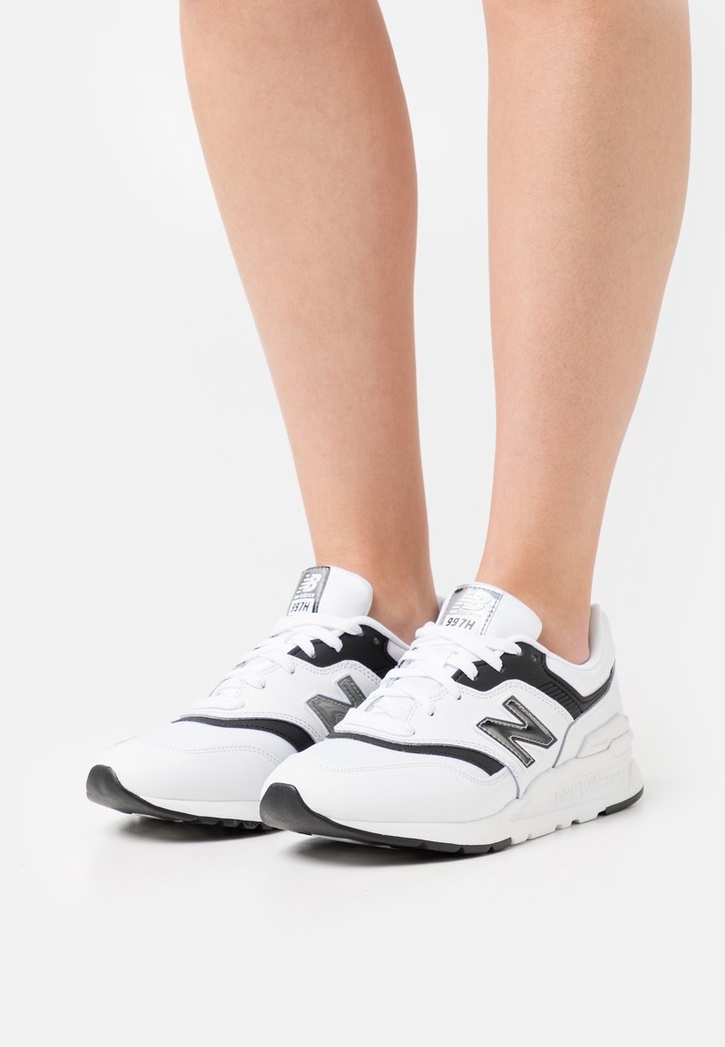 Osoba oblečená v bílých a černých teniskách New Balance 997H s tkaničkami, stojící na bílém pozadí.